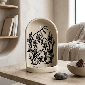 May include: Cream-colored ceramic wall decor with a black floral design. The arched piece features a detailed illustration of a plant with heart-shaped flowers and leaves. A small hole is at the top for hanging. The decor is on a light-colored table.