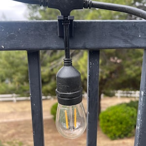 May include: A black outdoor string light with a clear bulb and a warm yellow filament is hanging from a black metal railing. The light is attached to a black wire and has a black plastic socket. The background is blurred with green foliage.