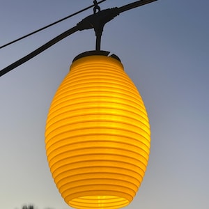May include: A bright yellow, oval-shaped paper lantern with horizontal ribbed detailing, suspended from a black electrical cord and wire system against a clear, light blue sky. The lantern emits a warm, glowing light from within its structure.