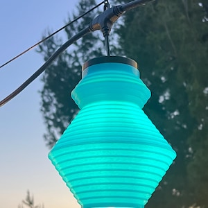 May include: A bright turquoise, ribbed, bell-shaped pendant light fixture hanging from a black electrical cord. The lamp features a distinct tiered silhouette with a wider base and a narrower top section, suspended against an outdoor sky.