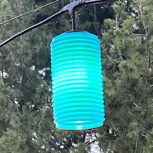 May include: A cylindrical, ribbed turquoise lantern light fixture suspended from a black electrical cord. The lantern features a translucent, horizontally ridged surface that glows with a bright, vibrant blue-green light, hanging outdoors.