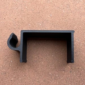 May include: A matte black 3D printed plastic clip featuring a C-shaped bracket design with a textured surface finish. The component includes an integrated circular hook attachment on the left side, designed for mounting or securing items.