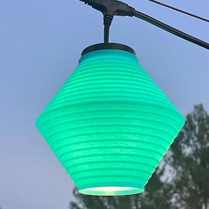 May include: A turquoise, diamond-shaped outdoor light fixture with horizontal ridges. The light hangs from a black wire and is set against a blue sky and tree background. The light is illuminated from within.
