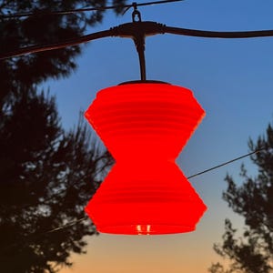 May include: A bright red, hourglass-shaped outdoor lantern hangs from a black wire against a twilight sky. The lantern has a ribbed texture and is suspended between trees. The background features a gradient of blue, orange, and yellow.