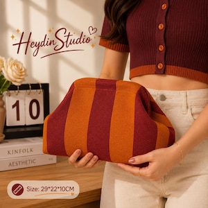 Könnte beinhalten: Eine gestreifte Clutch in Orange und Burgunderrot. Die Tasche hat vertikale Streifen und eine abgerundete Form. Die Maße sind 73 cm lang, 56 cm breit und 10 cm tief. Der Text "Heydin Studio" ist sichtbar.