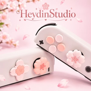 Pode incluir: Controlo de jogos branco com botões rosa e brancos e detalhes florais. O controlo tem uma cruz direcional preta e o texto "HeydinStudio" em uma fonte rosa elegante. O fundo é rosa suave com pétalas de flores de cerejeira.