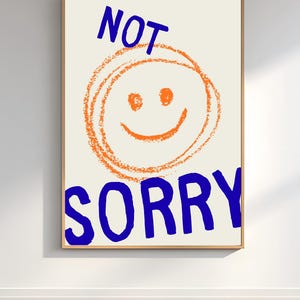 Puede incluir: Una impresión artística enmarcada con un fondo crema. La impresión presenta una gran carita sonriente naranja con las palabras "NOT" arriba y "SORRY" abajo, ambas en letras azules y en negrita. La obra de arte tiene una estética lúdica y moderna.