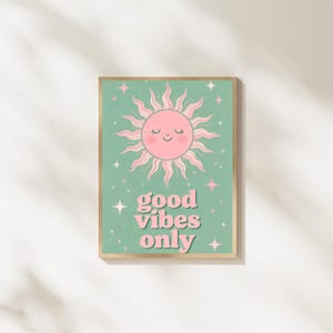 Op de afbeelding: Een ingelijste kunstdruk met een lachende roze zon en stralende lijnen op een zeegroene achtergrond. De woorden "good vibes only" staan in roze script onder de zon, met steraccenten. De lijst is goudkleurig.