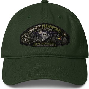 May include: A dark green baseball cap with a curved peak. The cap displays a patch with the text "800-WHO PARANORMAL RESEARCH SOCIETY" and a cartoon ghost cat. Additional text includes "Spectral Analyst" and "Bunk Industries".