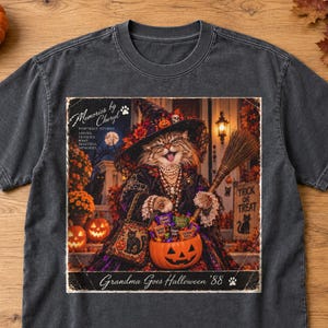 May include: A charcoal gray t-shirt featuring a graphic of a cat dressed as a witch, holding a broom and a pumpkin bucket filled with candy. The text reads "Memories by Cheryl," "Portrait Studio," "Loving Families Make Beautiful Memories," and "Grandma Goes Halloween '88."