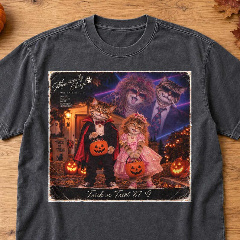 May include: A charcoal grey t-shirt featuring a graphic print of two cats dressed in Halloween costumes, one as a vampire and one in a pink dress, holding pumpkin buckets. The text reads Memories by Cheryl Portrait Studio and Trick or Treat '87.