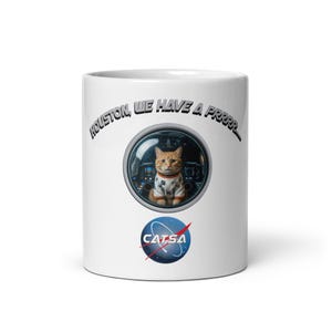 May include: A white ceramic mug featuring a graphic of an orange tabby cat in a space suit inside a circular window. Above the window is the text "HOUSTON, WE HAVE A PRRRR..." and below is a blue and white circular logo with the text "CATSA".
