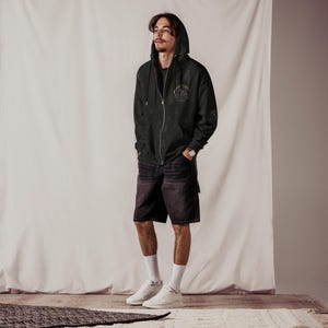 May include: A person wearing a black zip-up hoodie with a small graphic on the chest, black denim shorts, white socks, and white sneakers. The hoodie has a hood and a zipper closure. The shorts have pockets. The person is standing in front of a white backdrop.