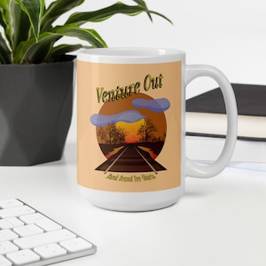May include: White ceramic mug featuring the text "Venture Out" above a graphic of a sunset over a railroad track. The mug has a large handle and is set against a neutral background.