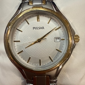 May include: A silver and gold-toned Pulsar watch with a white face and a date display at the 3 o'clock position. The watch features gold hands and markers, and a silver band. The watch face has the word "PULSAR" printed on it.