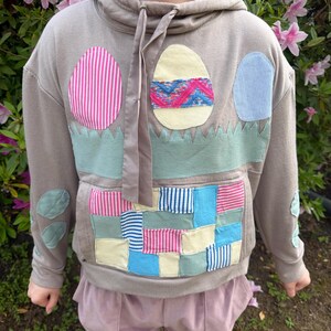 May include: A gray hoodie with Easter egg appliqués. The hoodie features a large front pocket and decorative patches in various colors and patterns. The Easter egg designs include pink stripes, blue stripes, and a woven design.