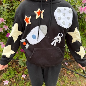May include: Black hooded sweatshirt with a space theme. The design includes a white rocket, a gray moon, an astronaut, and various stars in orange and cream colors. The sweatshirt has a front pocket and a drawstring hood.