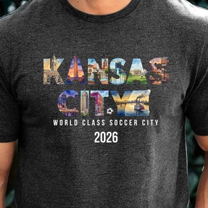 May include: A dark heather gray short-sleeve t-shirt featuring the text KANSAS CITY filled with colorful city landmark imagery. Below the text, the words WORLD CLASS SOCCER CITY and the year 2026 are printed in white, along with a small soccer ball icon.