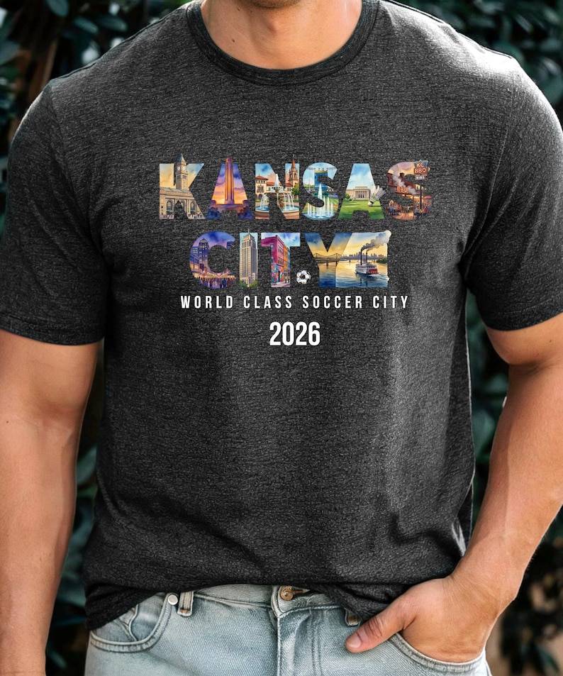 May include: A dark heather gray short-sleeve t-shirt featuring the text KANSAS CITY filled with colorful city landmark imagery. Below the text, the words WORLD CLASS SOCCER CITY and the year 2026 are printed in white, along with a small soccer ball icon.