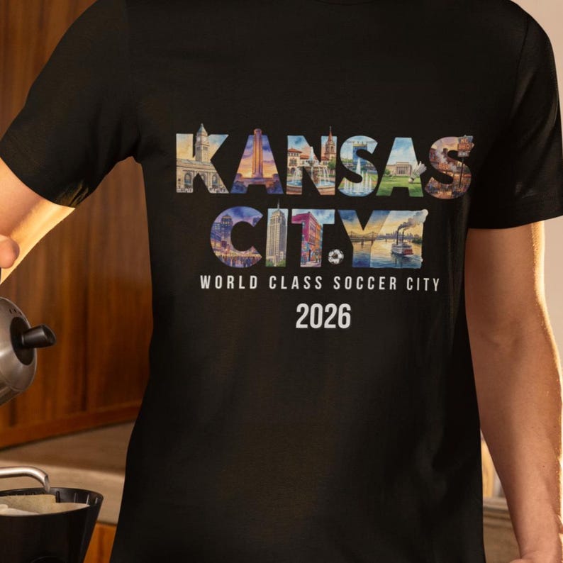 May include: A black t-shirt featuring the text KANSAS CITY in large letters filled with various city landmark images. Below this, the text reads WORLD CLASS SOCCER CITY and 2026 in white font. The design is centered on the front of the shirt.
