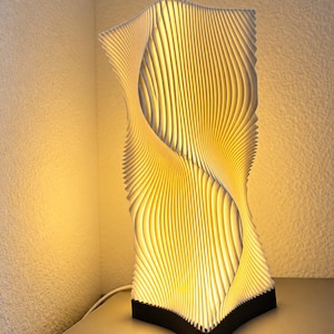 Puede incluir: Una lámpara de mesa vertical con una estructura blanca de múltiples capas en forma de onda que crea una silueta orgánica y retorcida. La lámpara está iluminada desde el interior, proyectando un brillo dorado cálido a través de los paneles.