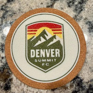 May include: Round cork coaster with a white center featuring the Denver Summit FC logo. The logo includes a mountain range, a rising sun, and the team's name in green and white. The coaster has a brown cork border.