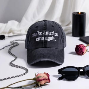 May include: A black baseball cap with the phrase "make america emo again" embroidered in white. The hat is distressed and sits on a white surface with a silver chain, sunglasses, and a rose.