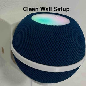 May include: A navy blue smart speaker with a white band, mounted on a white wall bracket. The speaker has a mesh-like texture and a glowing top with a multi-colored light. The text "Clean Wall Setup" is above the speaker.