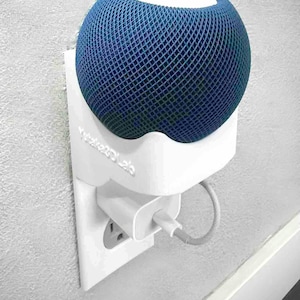 Könnte beinhalten: Ein blauer, mit Mesh bedeckter Smart-Lautsprecher befindet sich in einer weißen Wandhalterung, die an eine Steckdose angeschlossen ist. Der Halter ist mit "Wake2Dlabs" beschriftet. Der Lautsprecher hat eine weiße Oberseite und ein graues Ladekabel.
