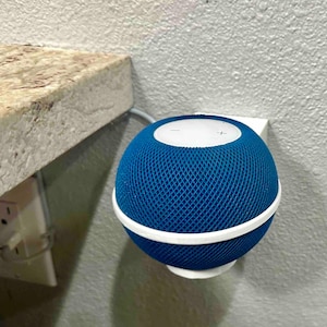 May include: A blue smart speaker with a white top and base, mounted on a wall. The speaker has a mesh fabric design and a minimalist aesthetic. The speaker is connected to a power outlet.