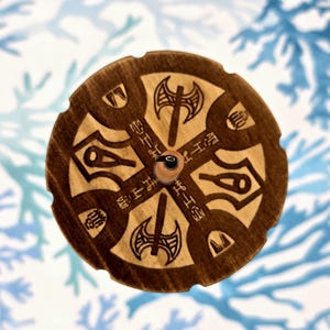May include: A round, wooden object with a dark brown finish. The design features etched axes, symbols, and Japanese characters arranged in a circular pattern. A small, black knob is in the center.