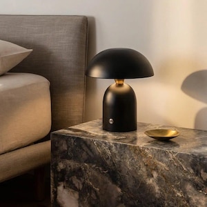 Peut inclure: Une lampe de table noire avec un abat-jour arrondi et une base dorée est posée sur une table d'appoint à motifs de marbre. Un petit bol doré est à côté de la lampe. La lampe est près d'un lit avec une tête de lit beige.