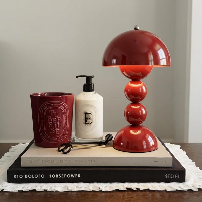 May include: A red table lamp with a dome-shaped shade and a stacked sphere design. Beside it are a red candle, a white soap dispenser, and a pair of black scissors, all arranged on books.