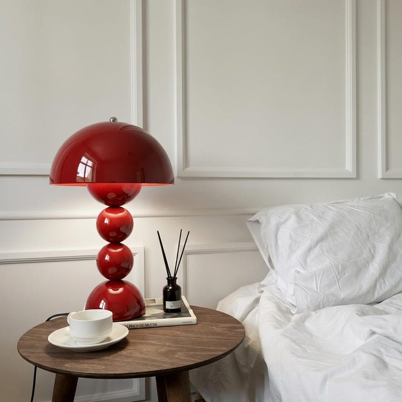 May include: A red table lamp with a rounded shade and stacked spherical base sits on a dark wood side table. A white teacup and saucer are on the table. The lamp is next to a bed with white linens.