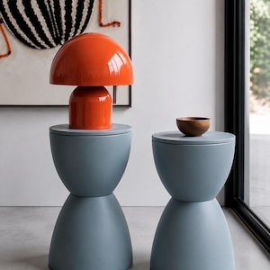 Pode incluir: Duas mesas laterais azul-claras em forma de ampulheta com superfícies lisas e foscas. Um candeeiro laranja brilhante em forma de cogumelo está na mesa da esquerda, enquanto uma pequena tigela redonda de madeira castanha natural está na direita.