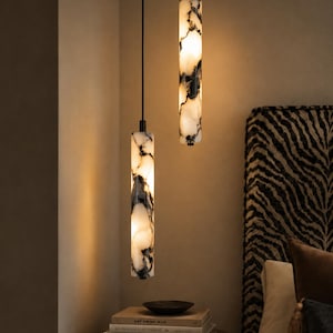 Pode incluir: Duas luminárias pendentes cilíndricas com um padrão de mármore branco e preto. Cada luminária é suspensa por um fio preto fino e emite um brilho quente a partir do interior. As peças têm um acabamento liso e polido em alturas diferentes.
