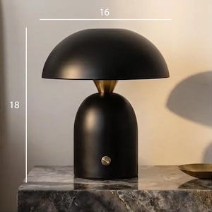 May include: A black table lamp with a rounded shade and base, accented with a brass-coloured neck. The lamp is 45.7 cm tall and 40.6 cm wide. The lamp has a single button on the base. The lamp is on a marble surface.