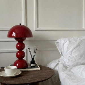 May include: A red table lamp with a spherical design, featuring a large dome-shaped shade and three stacked spheres. A white teacup and saucer sit on a dark wooden side table, next to a black diffuser and a book. A white bed is in the background.