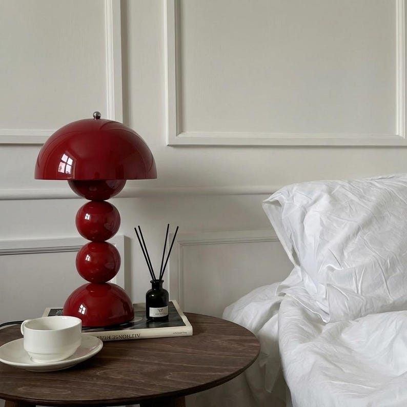 May include: A red table lamp with a spherical design, featuring a large dome-shaped shade and three stacked spheres. A white teacup and saucer sit on a dark wooden side table, next to a black diffuser and a book. A white bed is in the background.