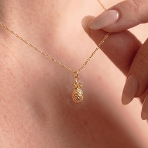 Op de afbeelding: Een goudkleurige ketting met een kleine ananas hanger. De hanger heeft een textuur en de ketting is delicaat. De ketting wordt om de nek gedragen.