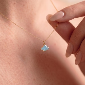Könnte beinhalten: Eine zarte goldene Halskette mit einem hellblauen Wolkenanhänger. Der Wolkenanhänger hat eine goldene Umrandung und hängt an einer feinen Goldkette. Die Halskette wird vor einem Hautton-Hintergrund gezeigt.