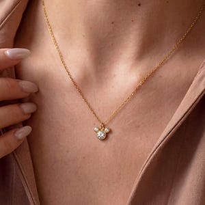 May include: A delicate gold necklace featuring a pendant with a three-stone design. The pendant has a central, larger clear stone, flanked by two smaller stones. The necklace is worn against a neutral background.