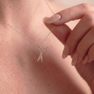 May include: A gold-toned necklace featuring a delicate chain and a small pendant shaped like a pair of scissors. The scissor blades are embellished with tiny, clear, sparkling stones, creating a refined and detailed jewelry piece.