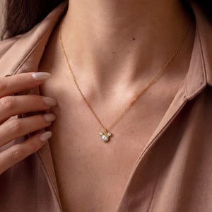 May include: A gold necklace with a pendant featuring three clear stones. The central stone is larger than the two flanking it. The necklace is suspended against a neutral background, highlighting the pendant's design.