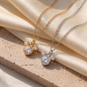 May include: Two necklaces with Mickey Mouse-shaped pendants. One is gold-coloured, the other silver-coloured, both featuring a large, round, clear gemstone centre and smaller side stones. The chains are fine, and the pendants are set against a cream-coloured fabric.