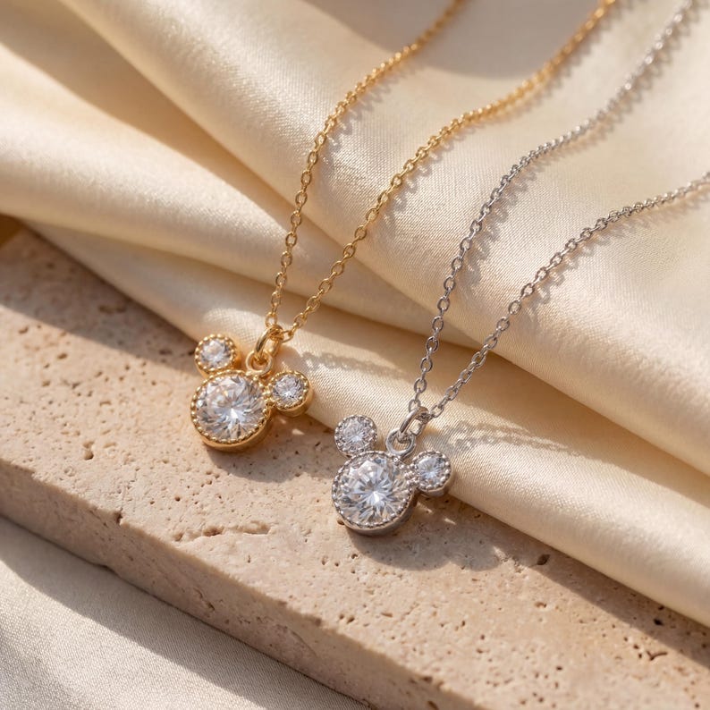 May include: Two necklaces with Mickey Mouse-shaped pendants. One is gold-coloured, the other silver-coloured, both featuring a large, round, clear gemstone centre and smaller side stones. The chains are fine, and the pendants are set against a cream-coloured fabric.