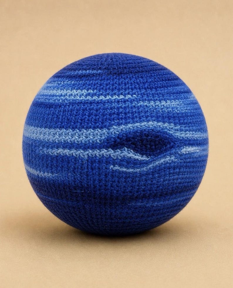 May include: A spherical object crafted with blue yarn, featuring a textured crochet stitch pattern. The surface displays horizontal bands of varying shades of blue, ranging from deep navy to lighter sky blue, creating a swirling, cloud-like effect.
