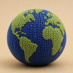 May include: A spherical, knitted globe featuring a deep blue background representing the oceans and textured, light green yarn detailing that forms the shapes of the continents. The surface has a consistent, visible knit stitch pattern throughout.