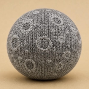May include: A spherical object made of grey knitted yarn, featuring a textured surface with lighter grey circular patterns that resemble craters on the moon. The knit construction is visible throughout the entire surface of the round item.