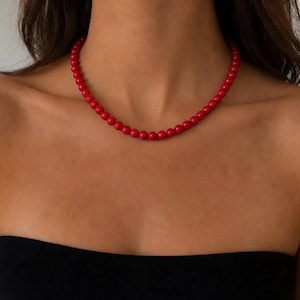 Peut inclure: Un collier à un seul rang composé de perles rouges vives, rondes, lisses et uniformes. Le collier repose près de la base du cou, formant une forme circulaire simple. Les perles ont une taille cohérente et une finition polie subtile.